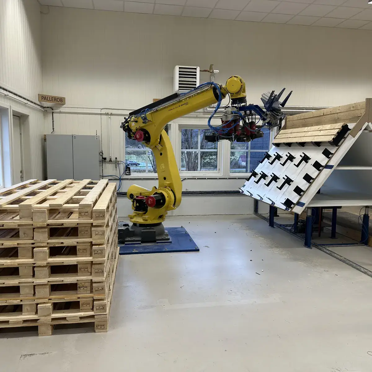 Gul industrirobot som setter sammen paller i SQPs produksjonshall, med en stabel ferdige trepaller i forgrunnen